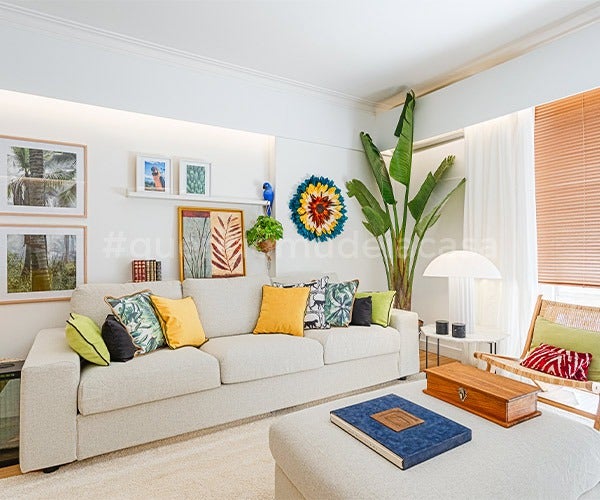 Sala com paredes brancas e decoração com cores vivas. Sofá com várias almofadas coloridas por cima, tapete bege e plantas