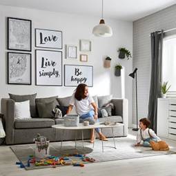 Mãe e filho em sala de estar, sentada em sofá, com parede de molduras, candeeiro suspenso branco e candeeiro de pé preto com tapete e almofadas para decorar