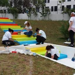 Grupo de pessoas a pintar umas escadas. Cada degrau tem uma cor